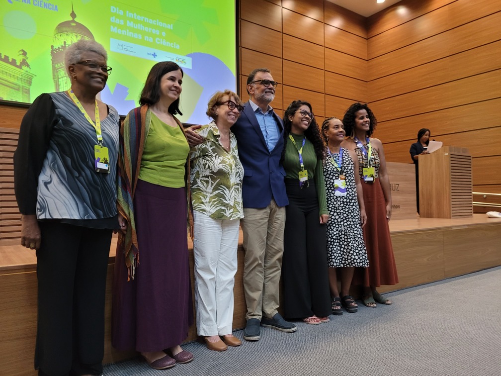 Foto de sete pessoas na frente do palco, no auditório, sendo seis mulhers e um homem.