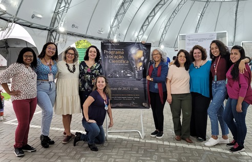 Equipe de pesquisadores e técnicos posando pra foto ao lado do banner da Semana de Vocação Cientifica do Provoc.