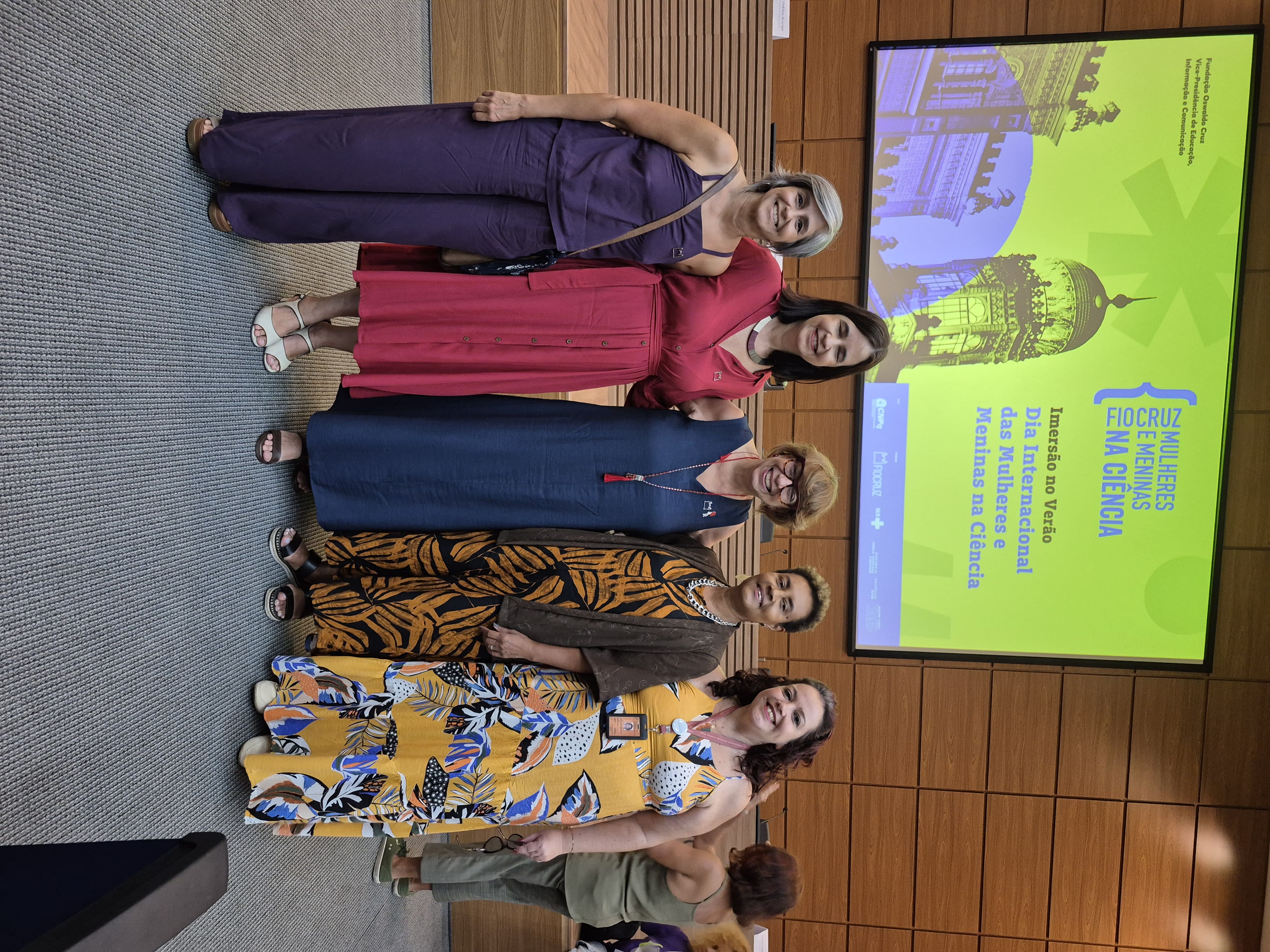 5 mulheres posam para foto na frente do painel do Mulheres e Meninas na Ciência.