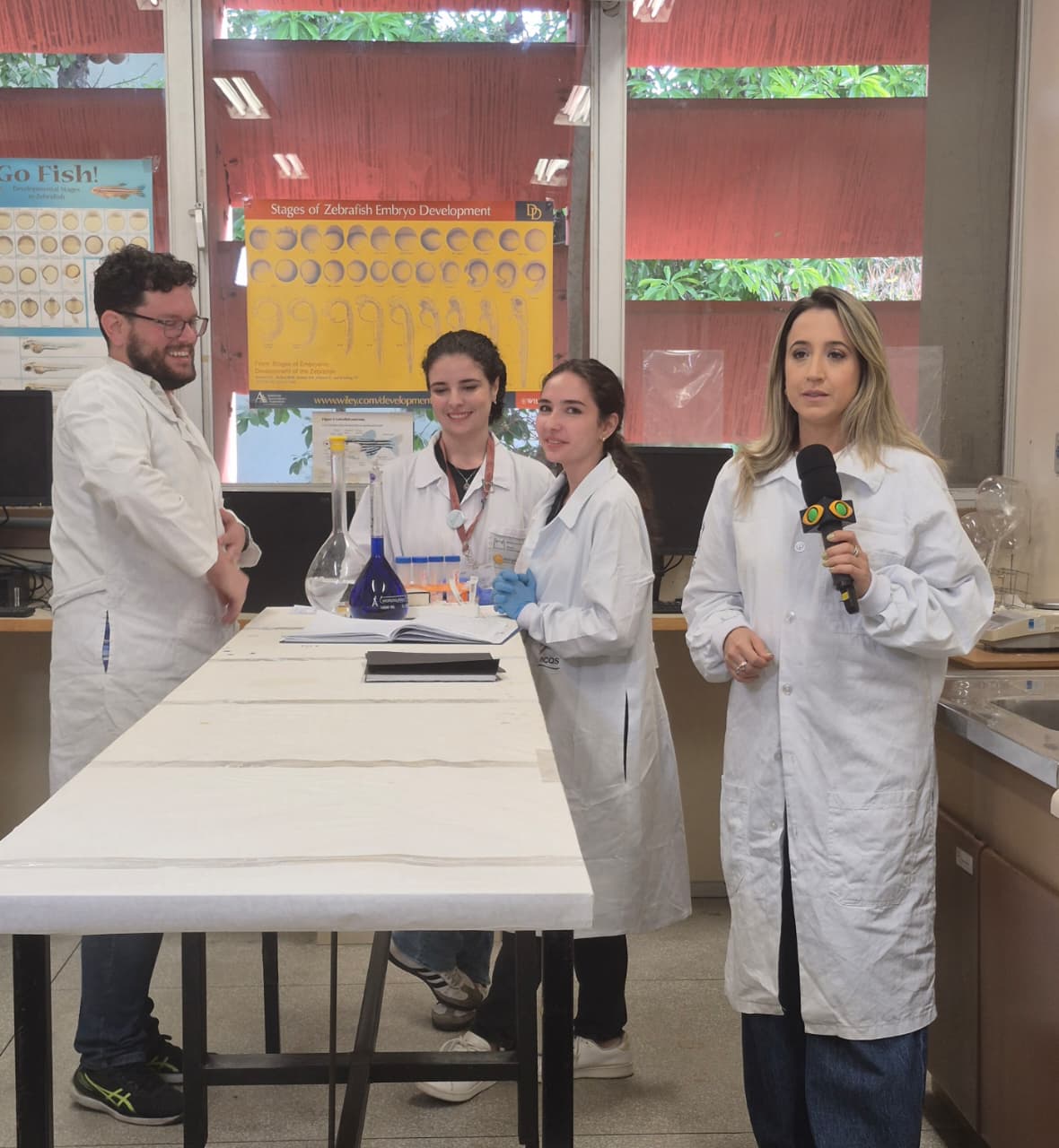 Repórter da Band TV fazendo a imagem da reportagem com as irmãs Juliana e Eliza Figueiredo e o orientador da Luiza ao fundo, no laboratório do INCQS da Fiocruz.