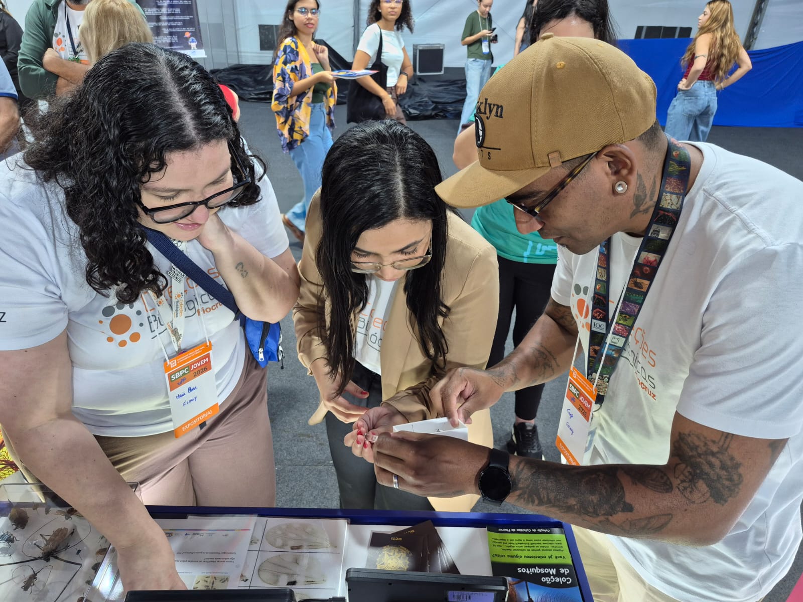 Três pessoas observam e manuseiam materiais de uma exposição científica em um estande durante evento de divulgação da ciência: SBPC Jovem.