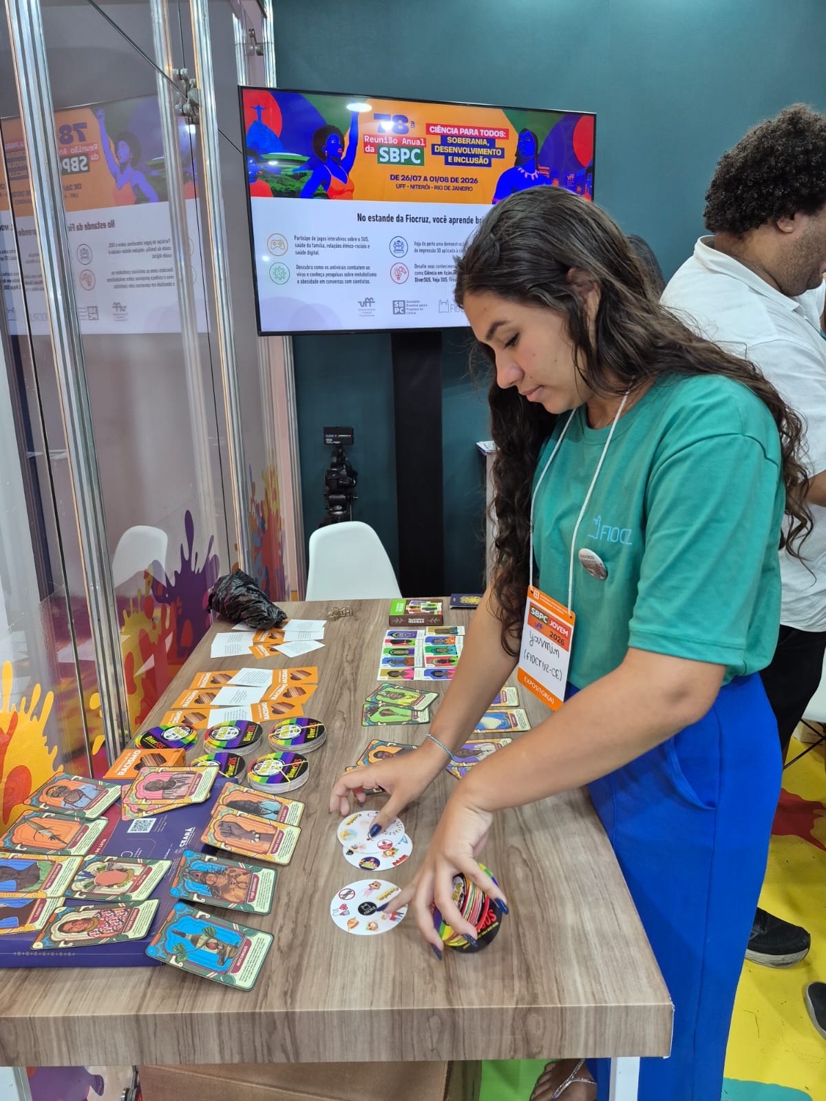 Pessoa organiza cartas e peças de um jogo educativo em uma mesa de exposição durante evento de divulgação científica. Ao fundo, há um monitor com informações da SBPC.