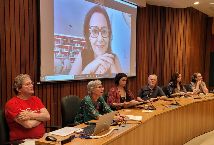 pessoas sentados atrás de uma mesa e uma tela com a participação de uma mulher.