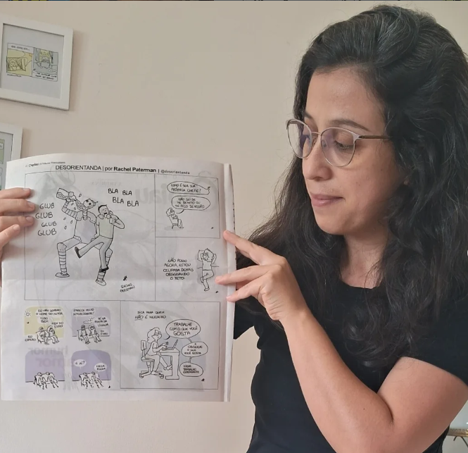 Pessoa com cabelos escuros e óculos está em pé diante de uma parede clara e segura uma folha com uma tirinha em quadrinhos. A pessoa aponta para a folha com uma das mãos. A tirinha tem desenhos em preto e branco e inclui o título “DESORIENTADA | por Rachel Paternan” no topo. Nos quadrinhos aparecem personagens com falas curtas em português, incluindo expressões como “blá blá blá” e “glub glub glub”. Ao fundo, na parede, há dois quadros pequenos com ilustrações. A iluminação é suave e natural, e a cena acontece em ambiente interno.