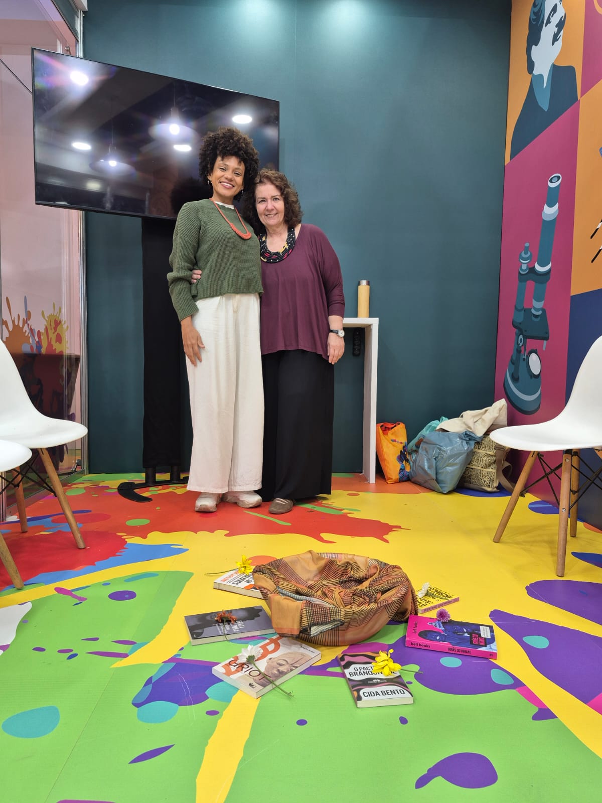 Duas pessoas posam juntas em uma sala colorida. No chão, há livros, flores e um tecido disposto em círculo. Ao fundo, vê-se uma TV desligada e um painel com ilustração de microscópio.