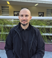 Pessoa careca, com barba curta, enquadrada do peito para cima, olhando para a câmera. A pessoa veste uma blusa escura com zíper e um casaco preto por cima. Ao fundo há um prédio com parede em tom rosado na parte superior e estrutura com cobertura metálica clara. Sob a cobertura, aparecem luminárias acesas e janelas com molduras claras. Na altura do peito da pessoa, há um corrimão metálico e, logo atrás, um canteiro com plantas verdes alongadas. 