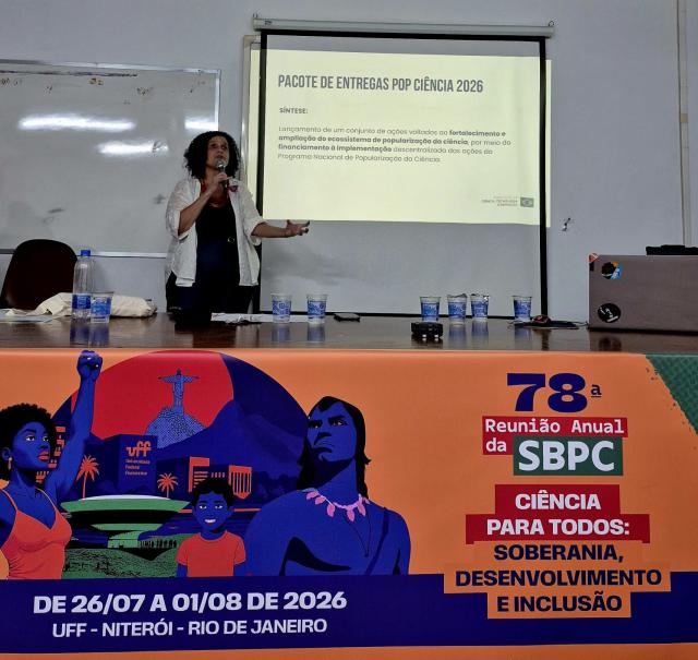 Pessoa apresenta uma palestra em auditório, falando ao microfone diante de uma projeção sobre o programa “Pacote de Entregas Pop Ciência 2026”. Na frente da mesa, há um painel da 78ª Reunião Anual da SBPC.