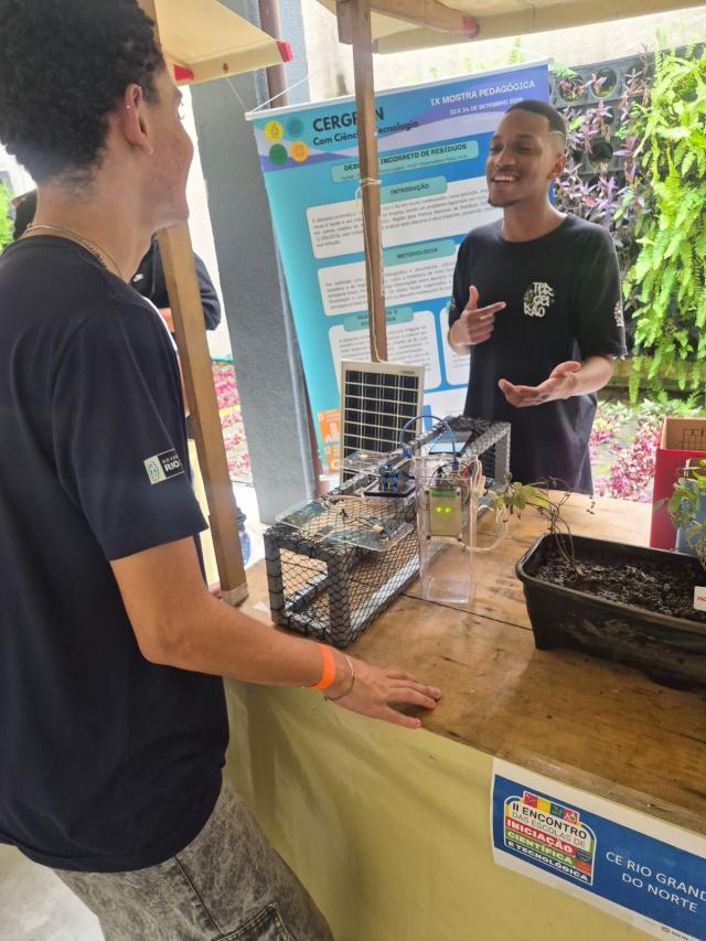 Estudante demonstra sistema automatizado para plantas a visitantes em estande de feira científica.