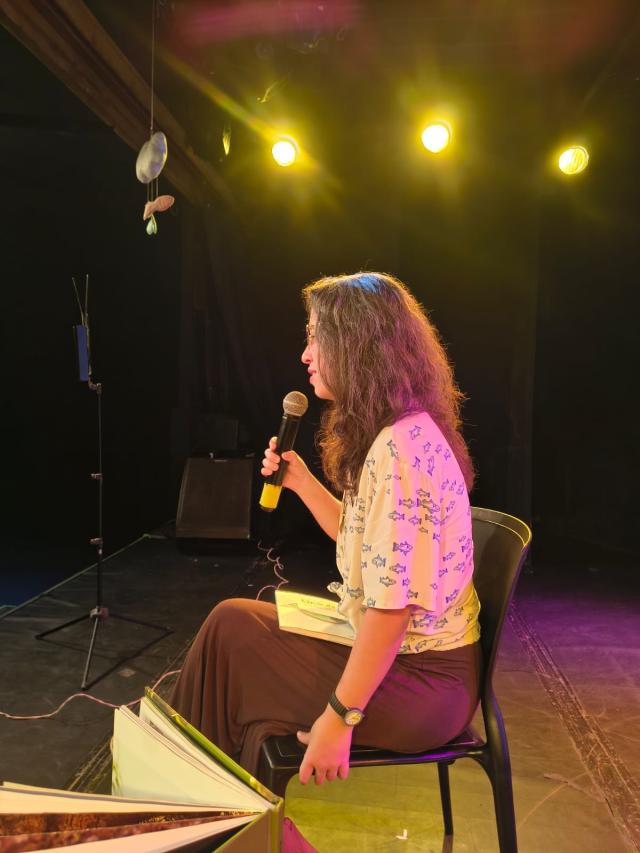 Pessoa sentada no palco segura um microfone e um livro no colo, iluminada por luzes amarelas, com um livro grande aberto à frente.