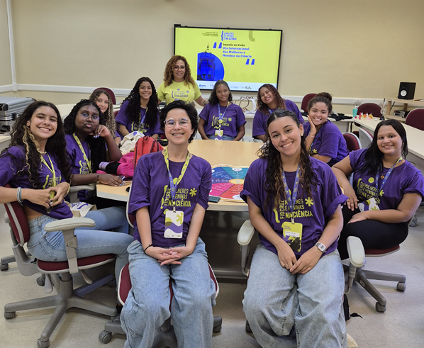 dez meninas em volta de uma mesa de reunião posam com a pesquisadora para foto.