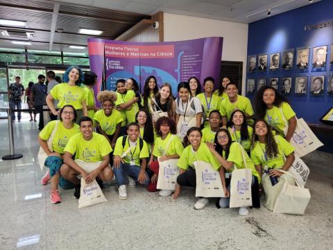 Foto de estudantes e professores sorrindo na frente do poster do evento Mulheres e Meninas na Ciência.