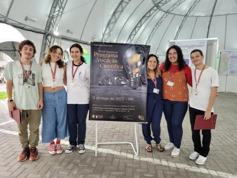 Seis estudantes da Etapa Avançado do Programa de Vocação Científica posam ao lado do cartaz do evento na Tenda da Escola Politécnica