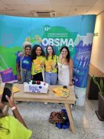 4 jovens do gênero feminino ganharam o jogo de perguntas e respostas de conhecimento cultural e científico posam felizes com seus brindes, na frente do cartaz da Olimpíada Brasileiras de Saúde e Meio Ambiente.