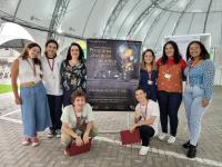 Seis estudantes da Etapa Avançado do Programa de Vocação Científica posam ao lado do cartaz do evento na Tenda da Escola Politécnica