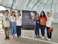 Seis estudantes da Etapa Avançado do Programa de Vocação Científica posam ao lado do cartaz do evento na Tenda da Escola Politécnica