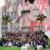 Grande grupo de estudantes do ensino médio, de diferentes estados do Brasil, posa sorrindo e com os braços erguidos em frente ao prédio da Escola Politécnica de Saúde Joaquim Venâncio, da Fiocruz. Ao fundo, há um mural com pintura de trabalhadores e elementos relacionados à saúde e à ciência.